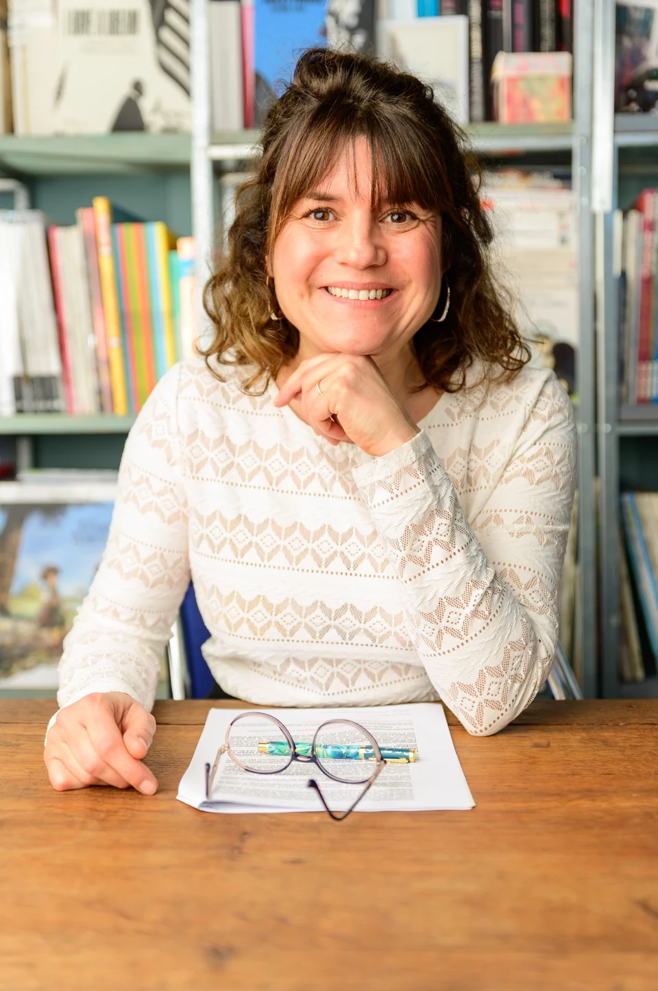 Julia Peironet-Brémond en haut clair devant sa bibliothèque, avec des documents et des lunettes sur la table